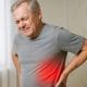 Hombre con dolor lumbar tocándose la zona baja de la espalda, con la región afectada resaltada en rojo en una clínica médica.