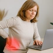 Mujer trabajando desde casa sentada con mala postura, tocándose la zona lumbar con signos de dolor, representando los efectos del sedentarismo en la espalda baja.